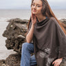 Woman sitting on a rock by the sea wearing a brown poncho with floral patterns.