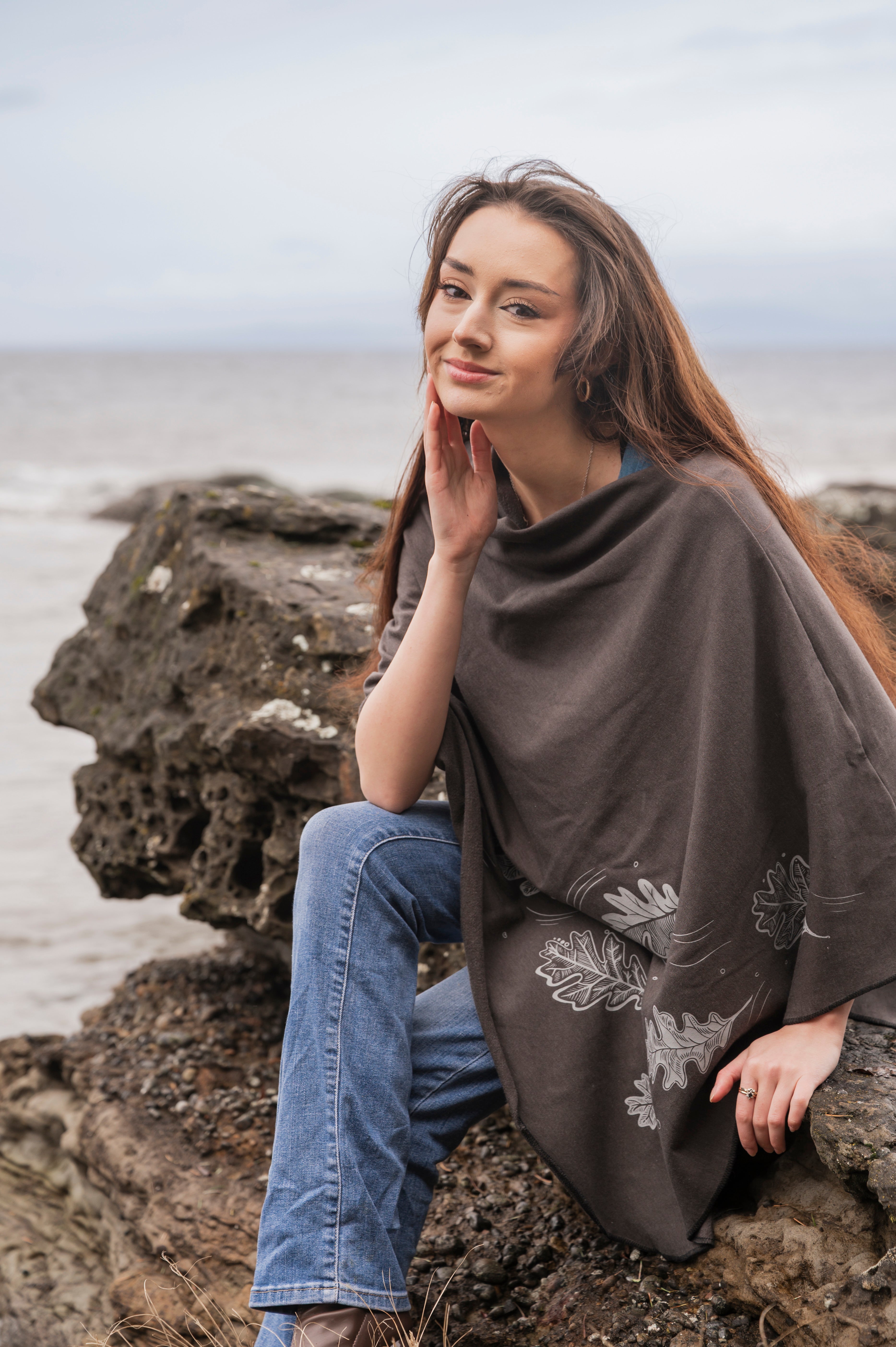 Woman sitting on a rock by the sea wearing a brown poncho with floral patterns.