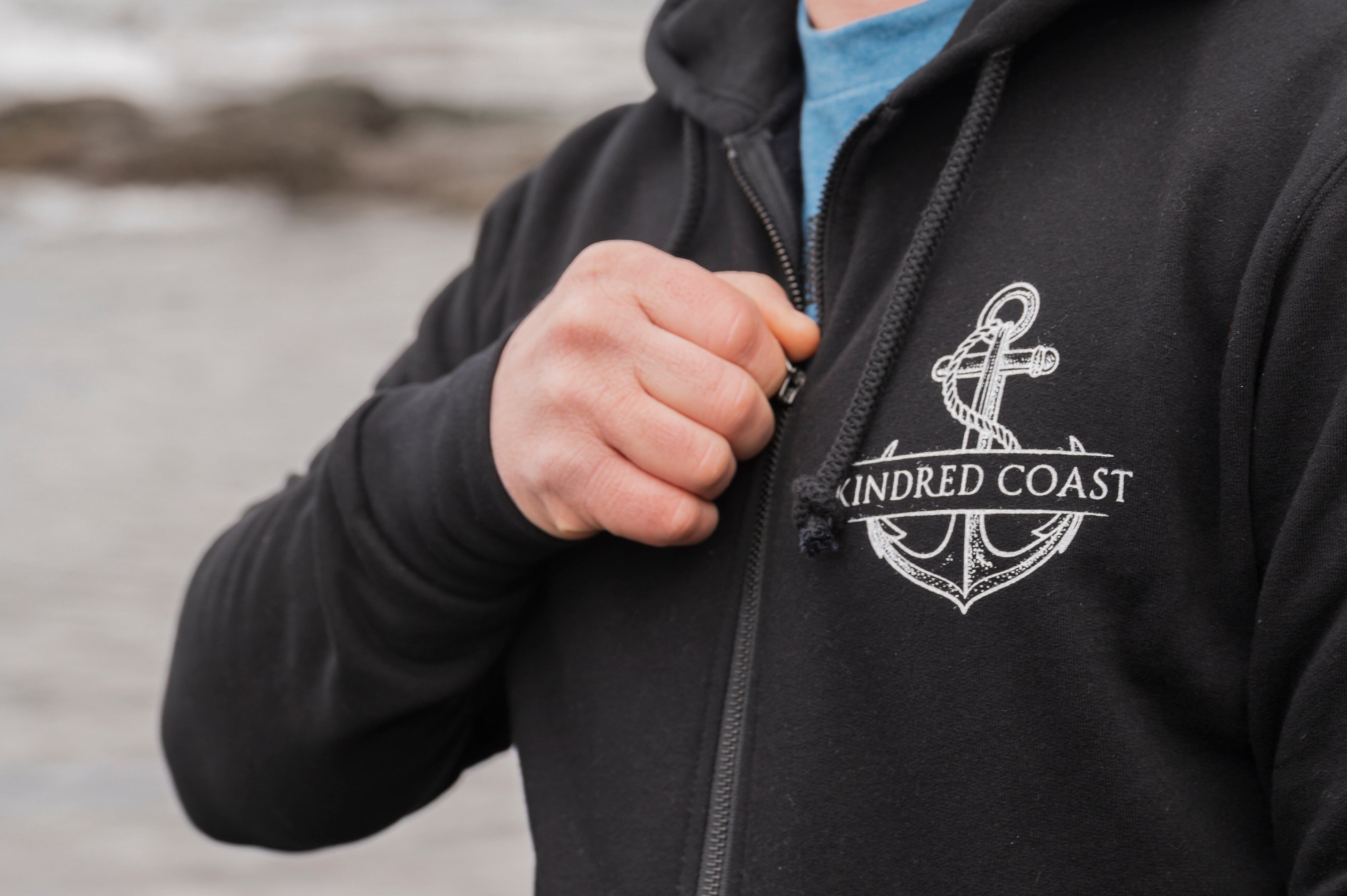 Man wearing a black hoodie with an anchor logo and 'Kindred Coast' text by the ocean.