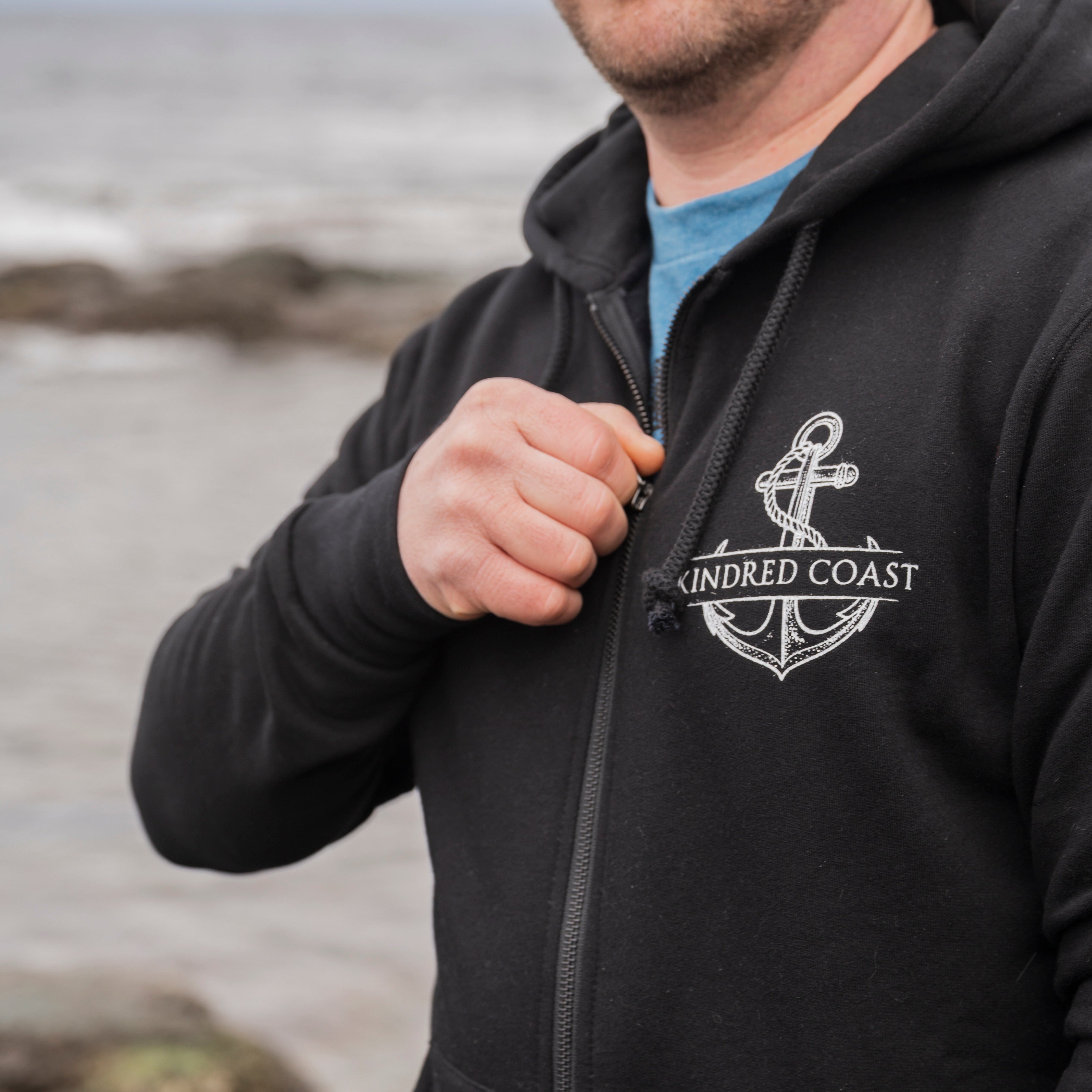 Man wearing a black hoodie with an anchor logo and 'Kindred Coast' text by the ocean.