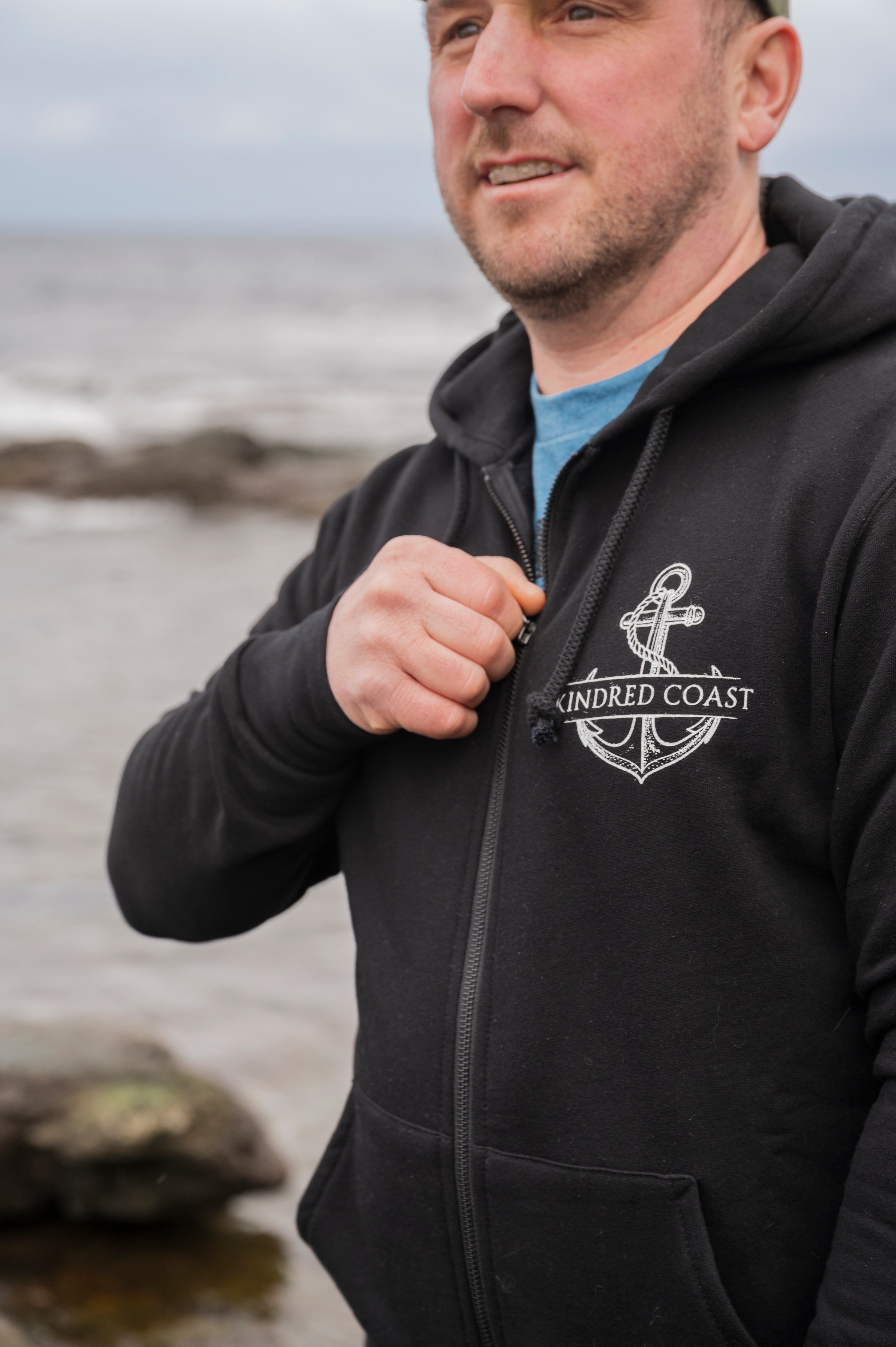 Man wearing a black hoodie with an anchor logo and 'Kindred Coast' text by the ocean.