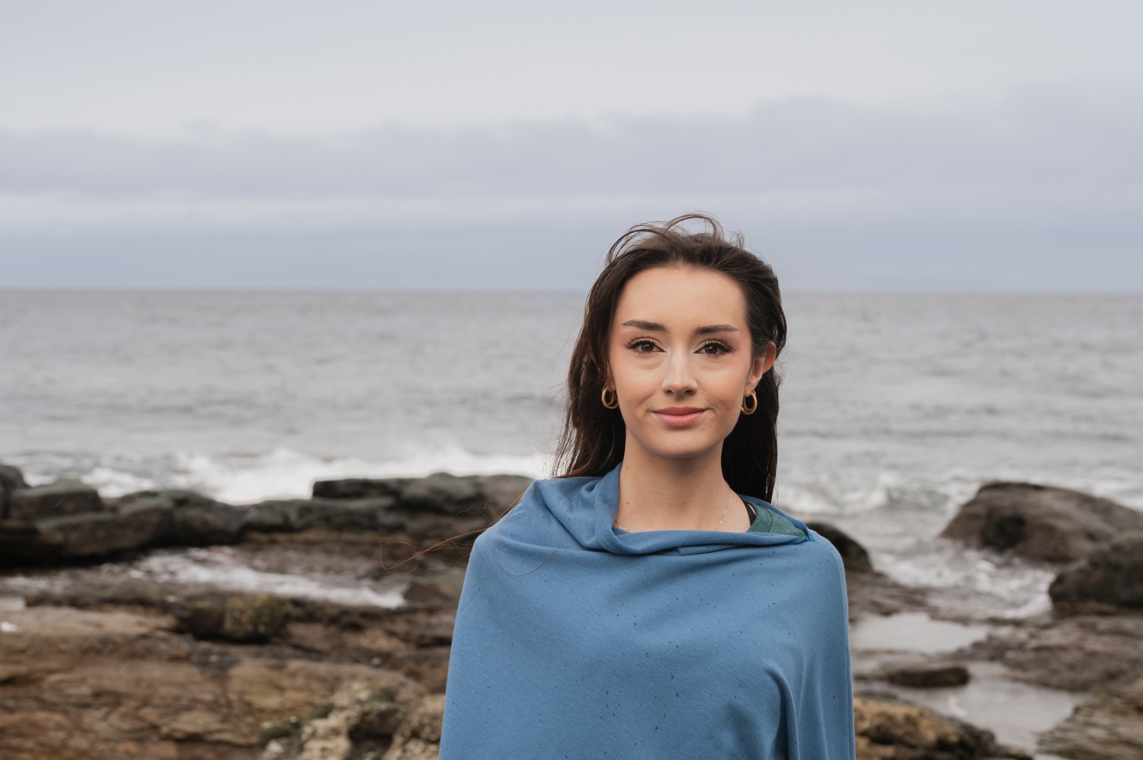 Woman wearing a blue poncho with floral patterns standing on a rocky beach.