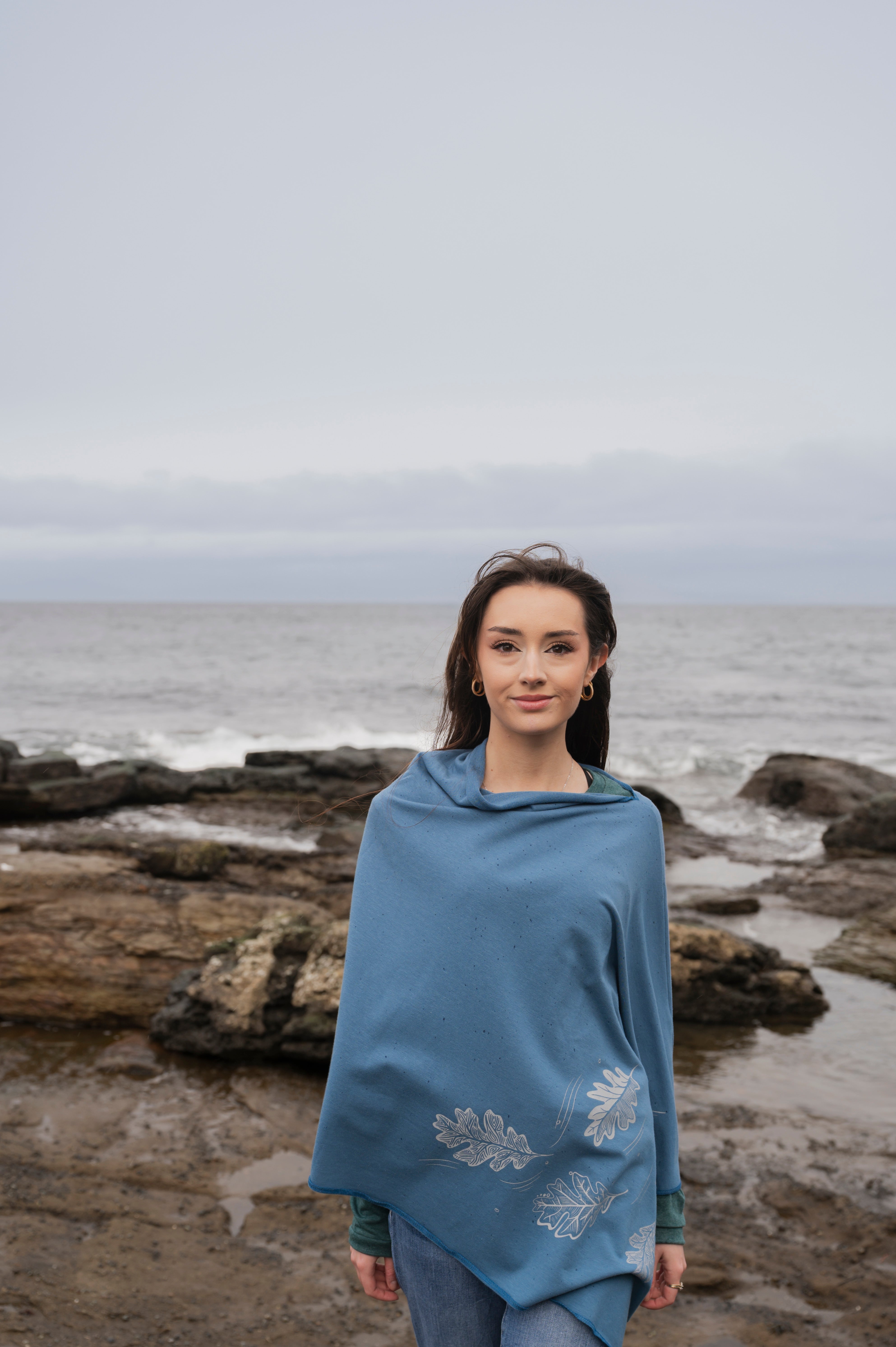 Woman wearing a blue poncho with floral patterns standing on a rocky beach.