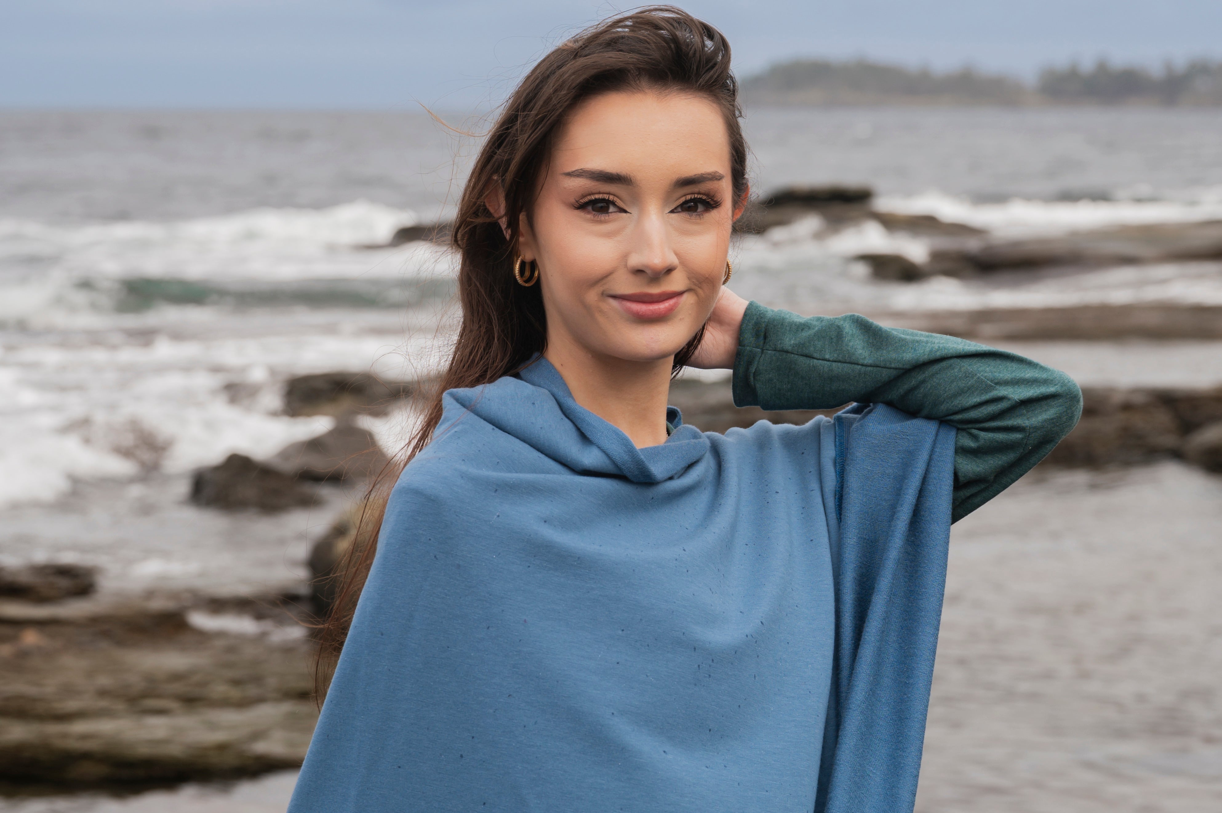 Woman wearing a blue poncho standing on a beach with ocean and driftwood in the background