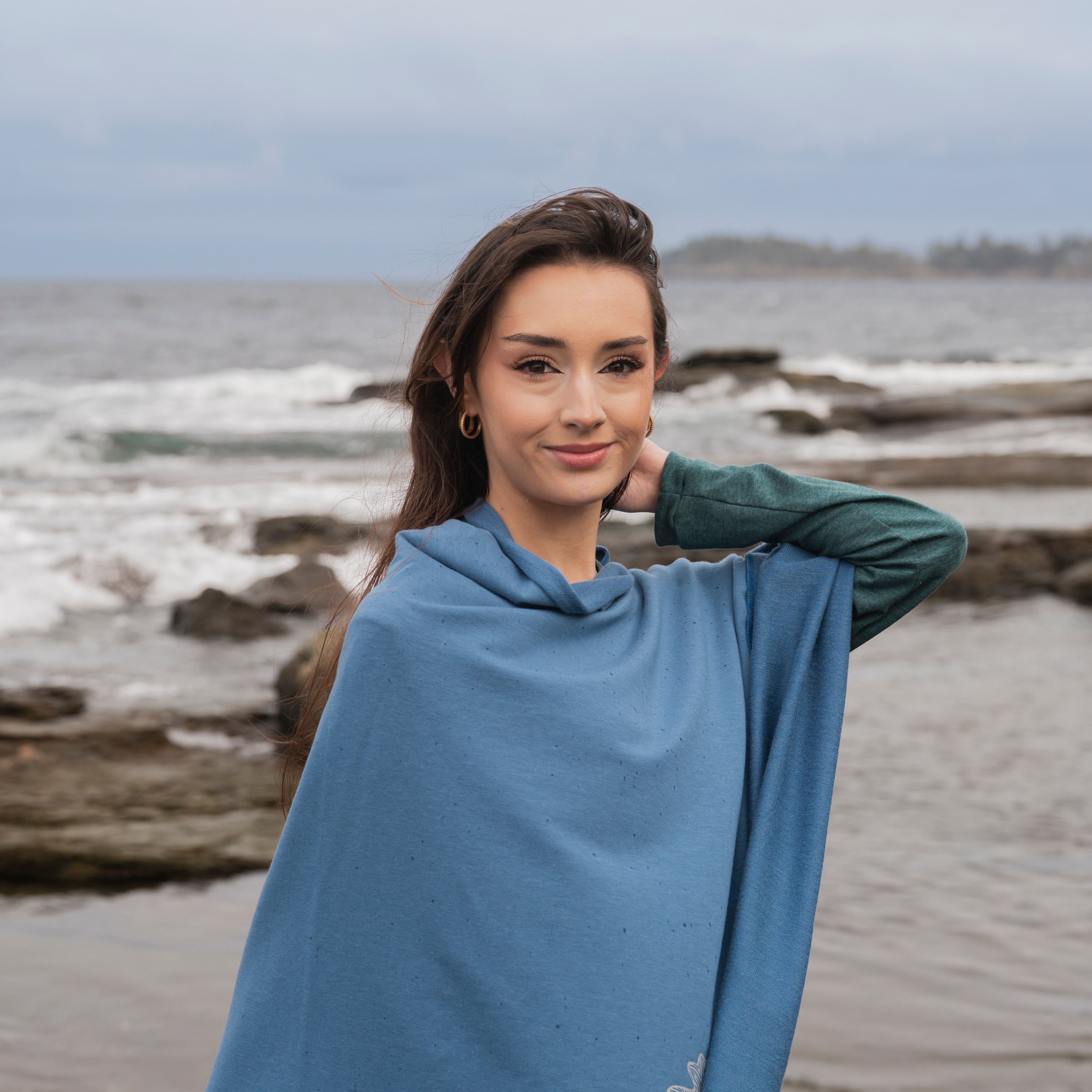 Woman wearing a blue poncho standing on a beach with ocean and driftwood in the background