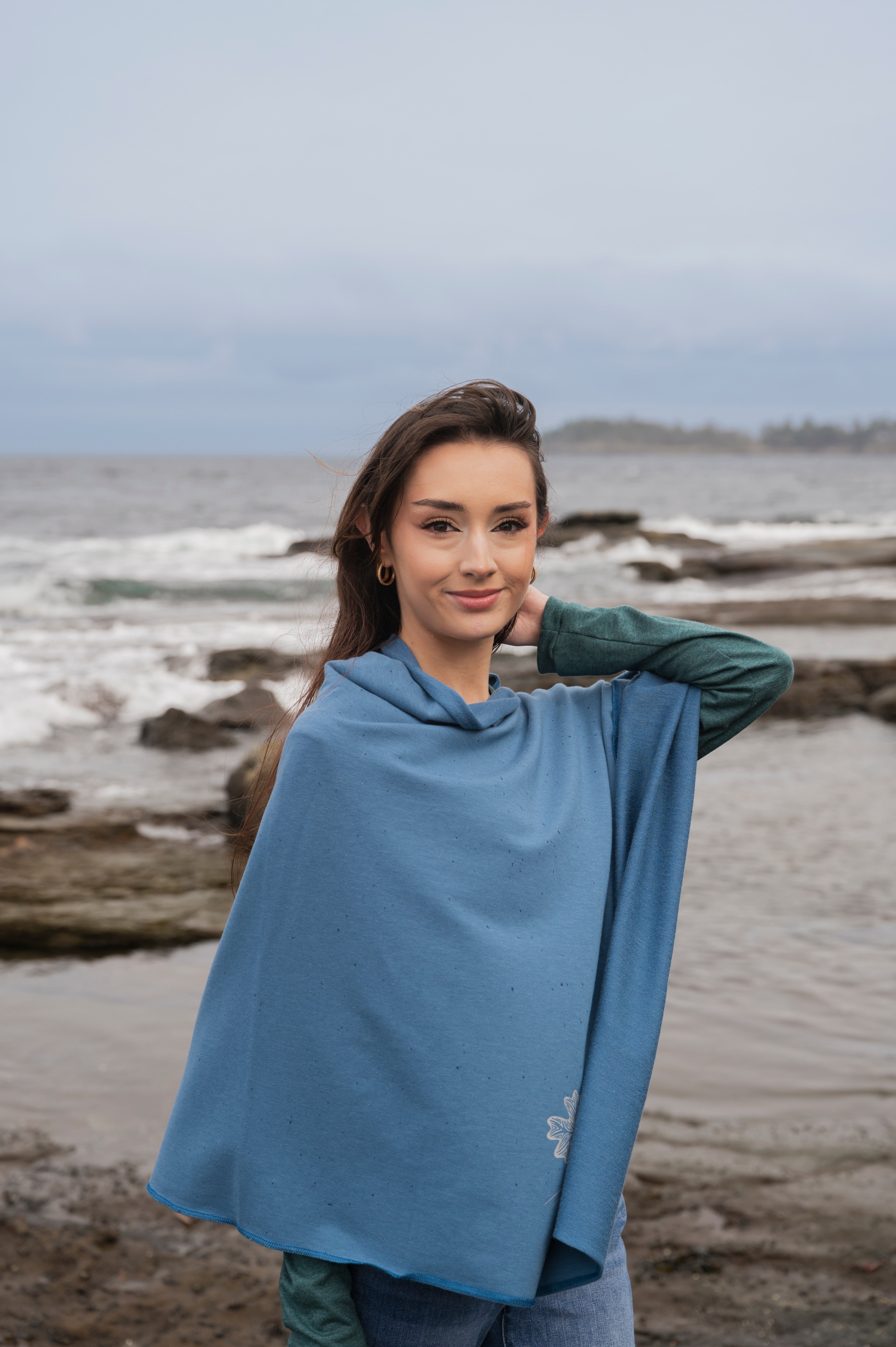 Woman wearing a blue poncho standing on a beach with ocean and driftwood in the background