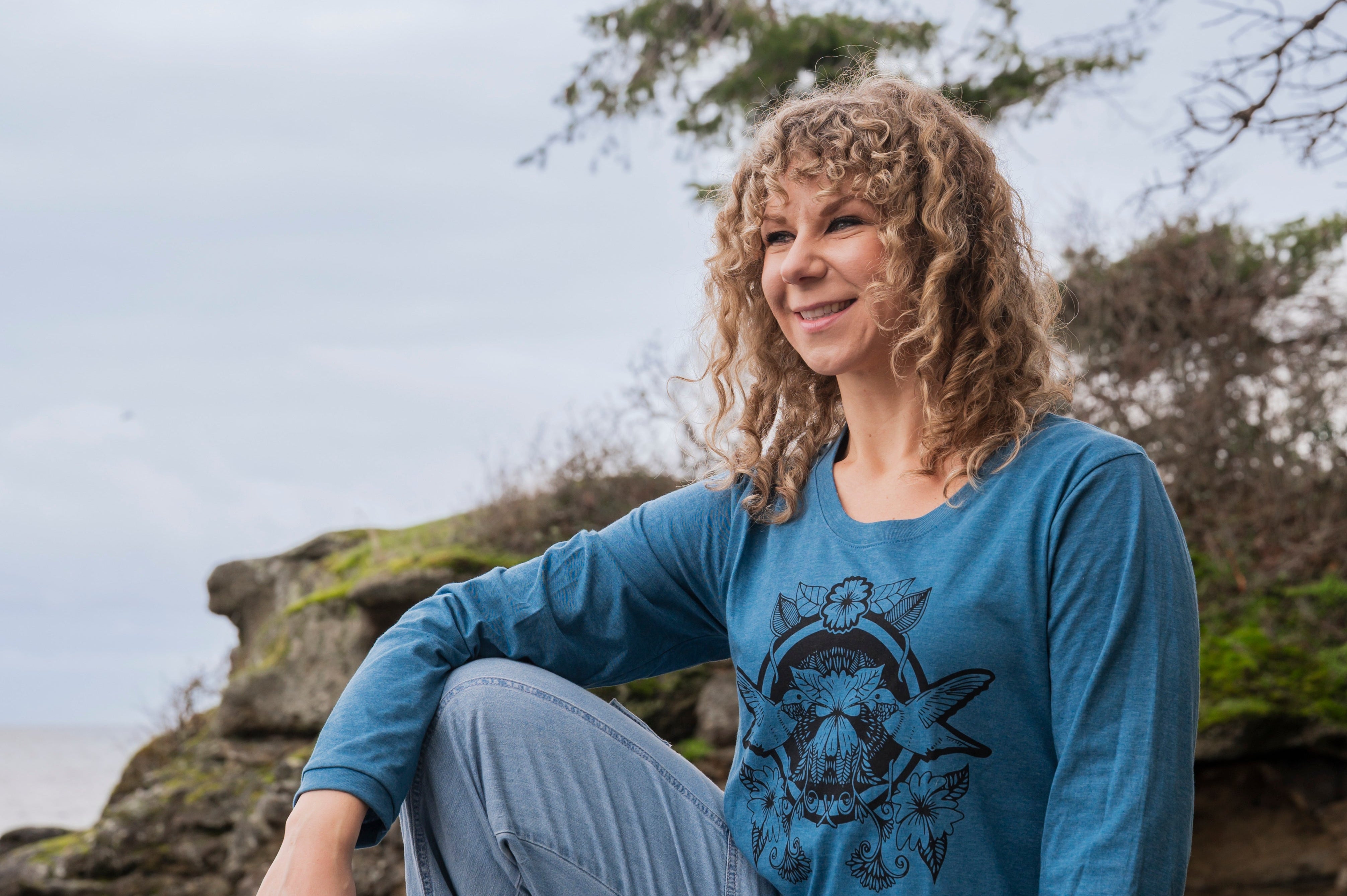 Woman sitting on a rock by the ocean wearing a blue long-sleeve shirt with a graphic design.