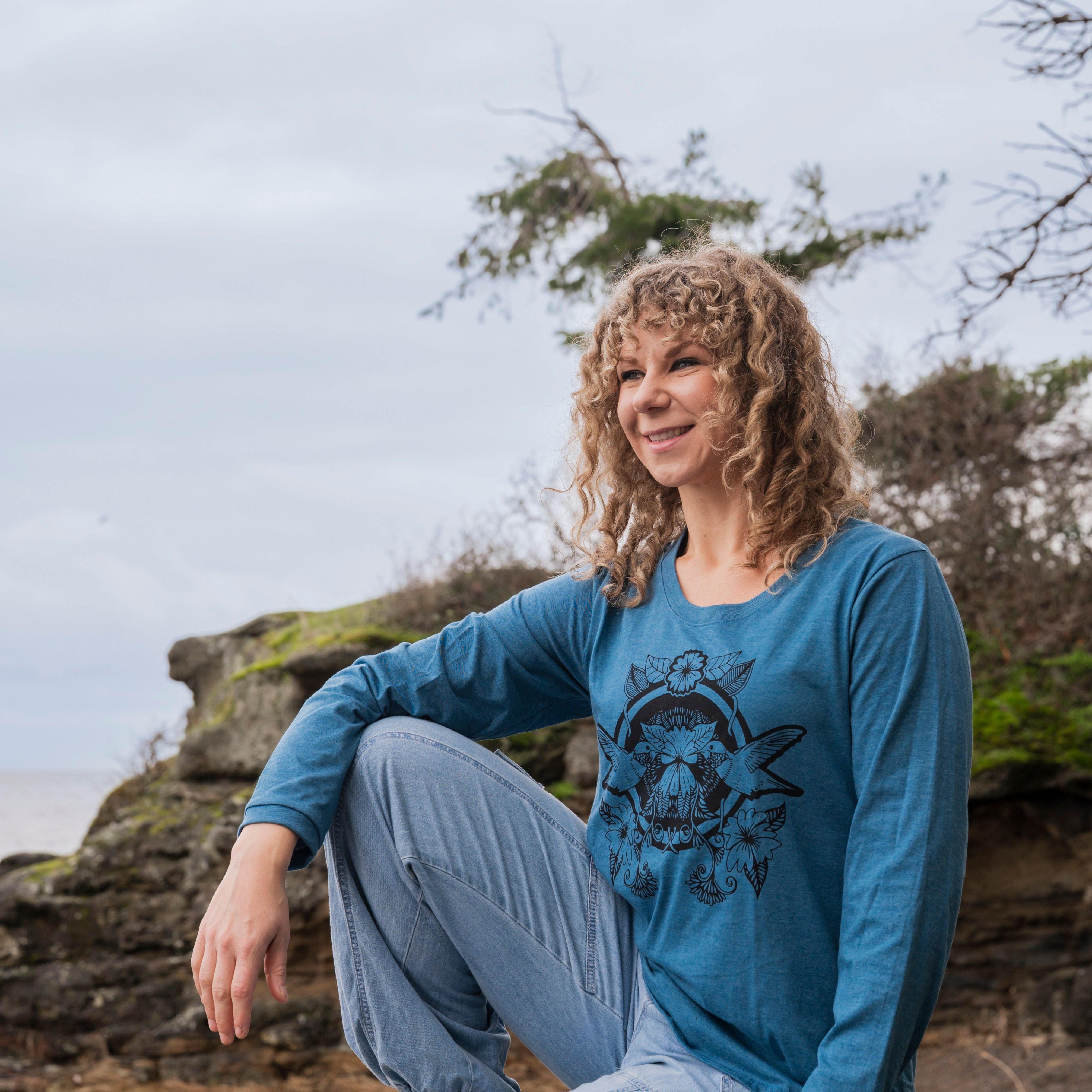 Woman sitting on a rock by the ocean wearing a blue long-sleeve shirt with a graphic design.