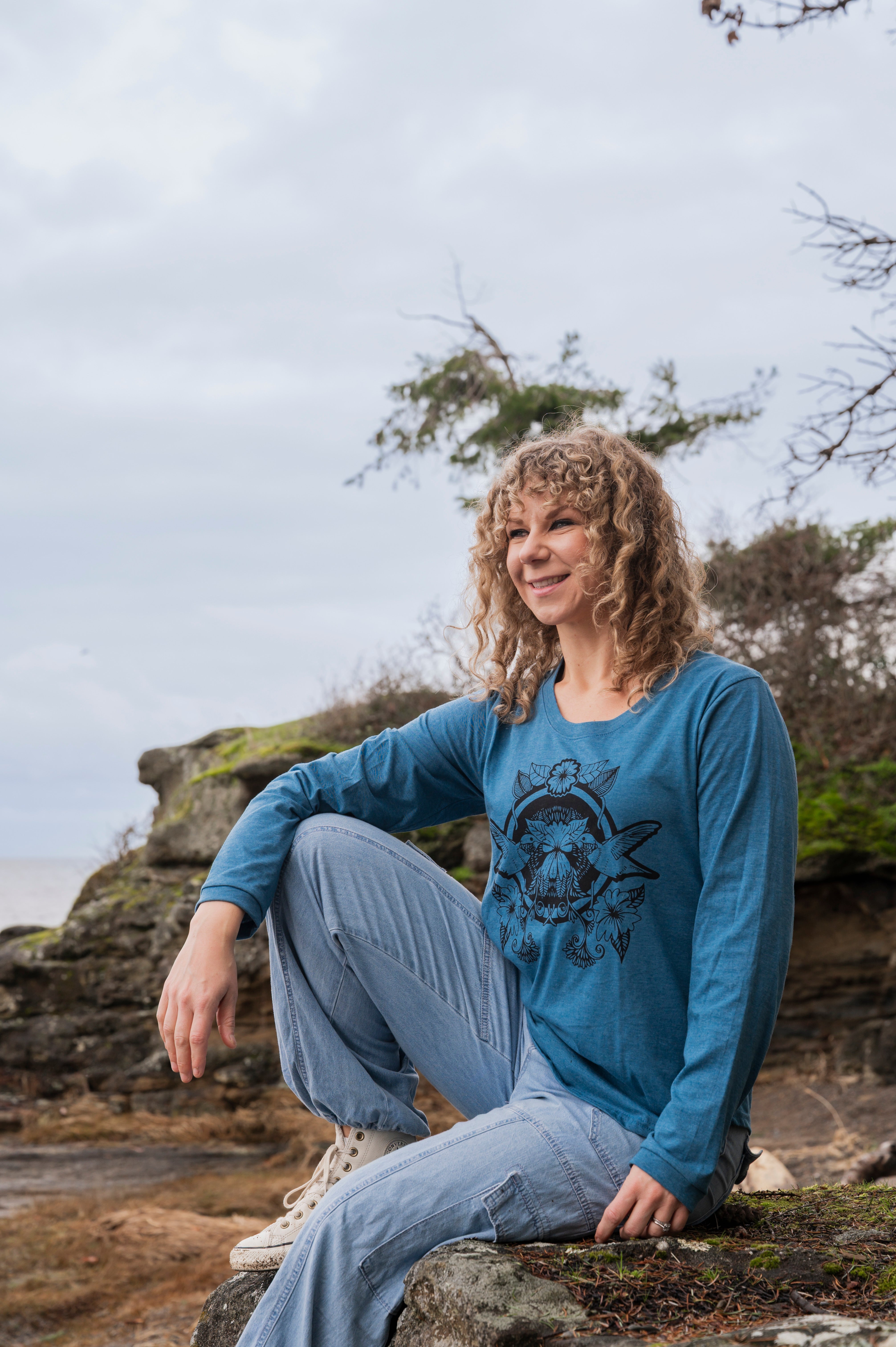 Woman sitting on a rock by the ocean wearing a blue long-sleeve shirt with a graphic design.