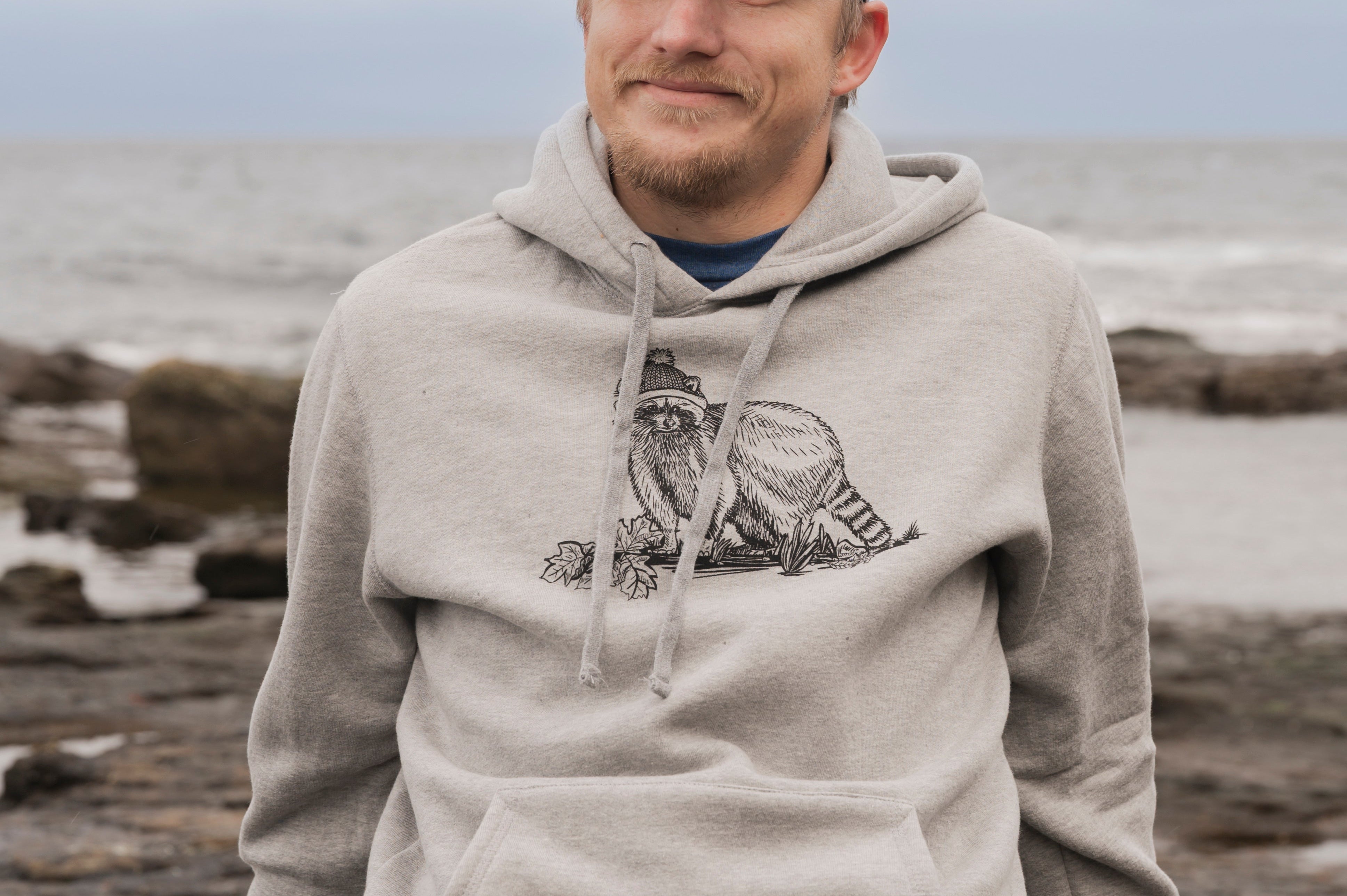 Man wearing a gray hoodie with a logo, sitting on rocks by the ocean.