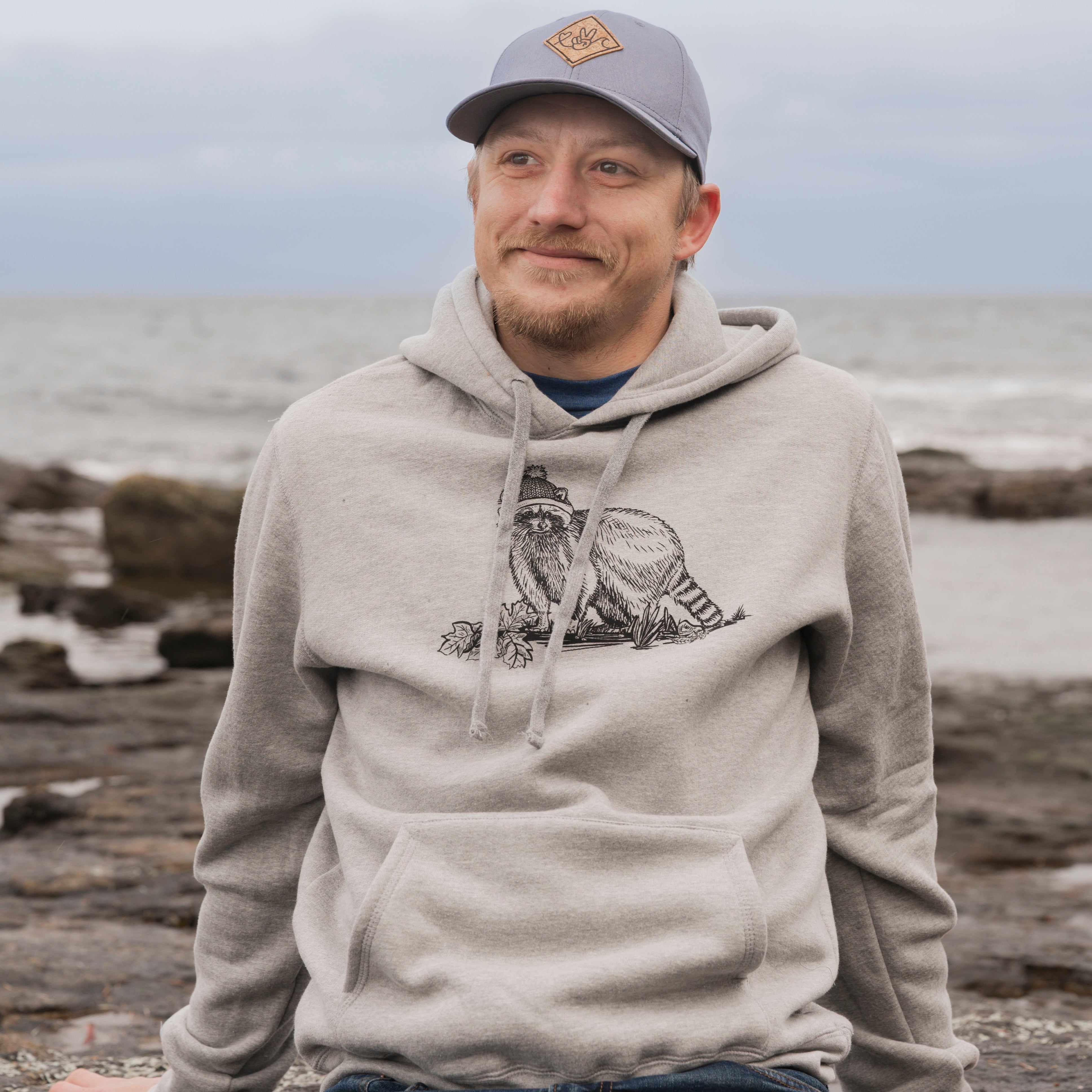 Man wearing a gray hoodie with a logo, sitting on rocks by the ocean.