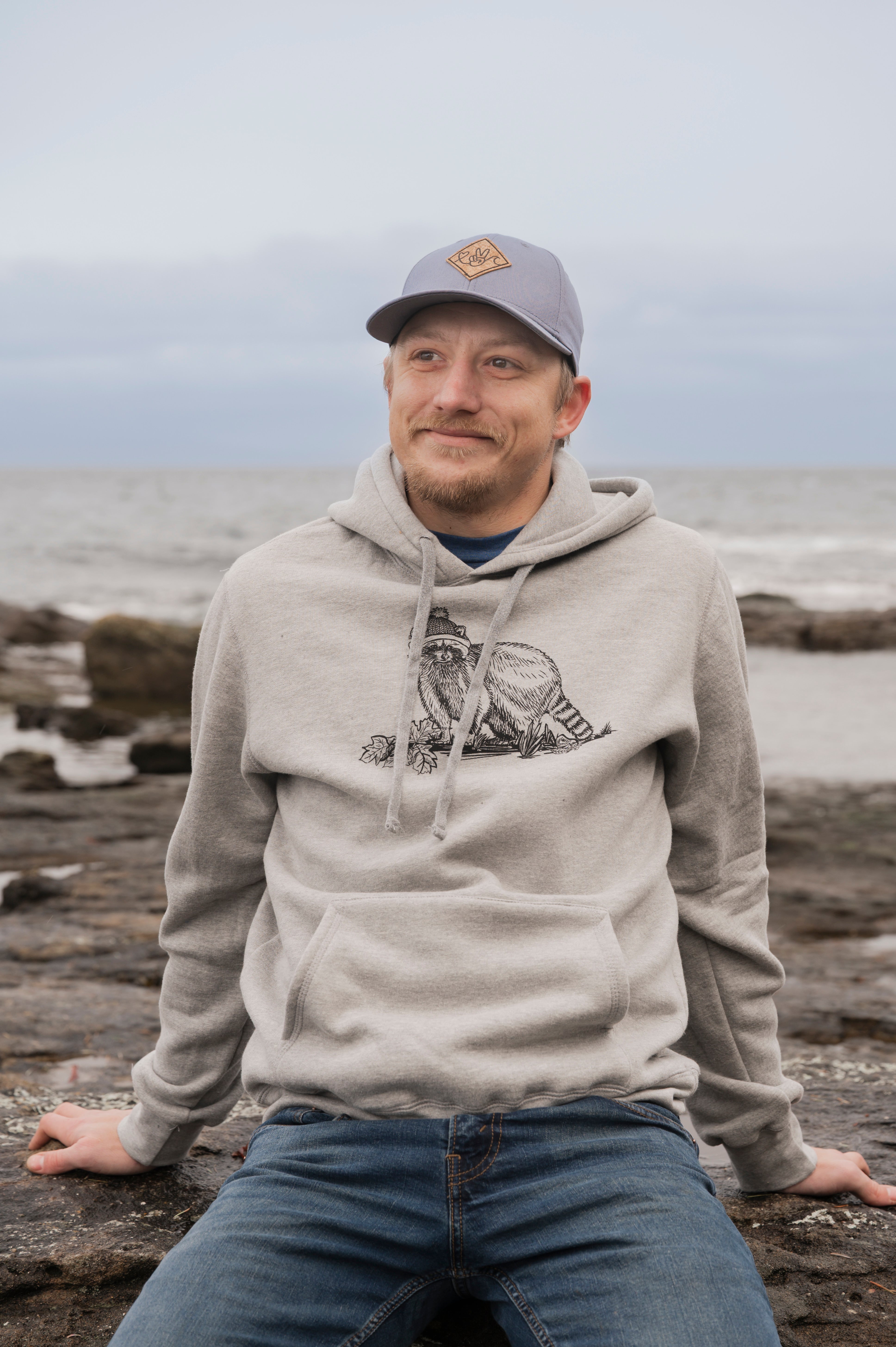 Man wearing a gray hoodie with a logo, sitting on rocks by the ocean.