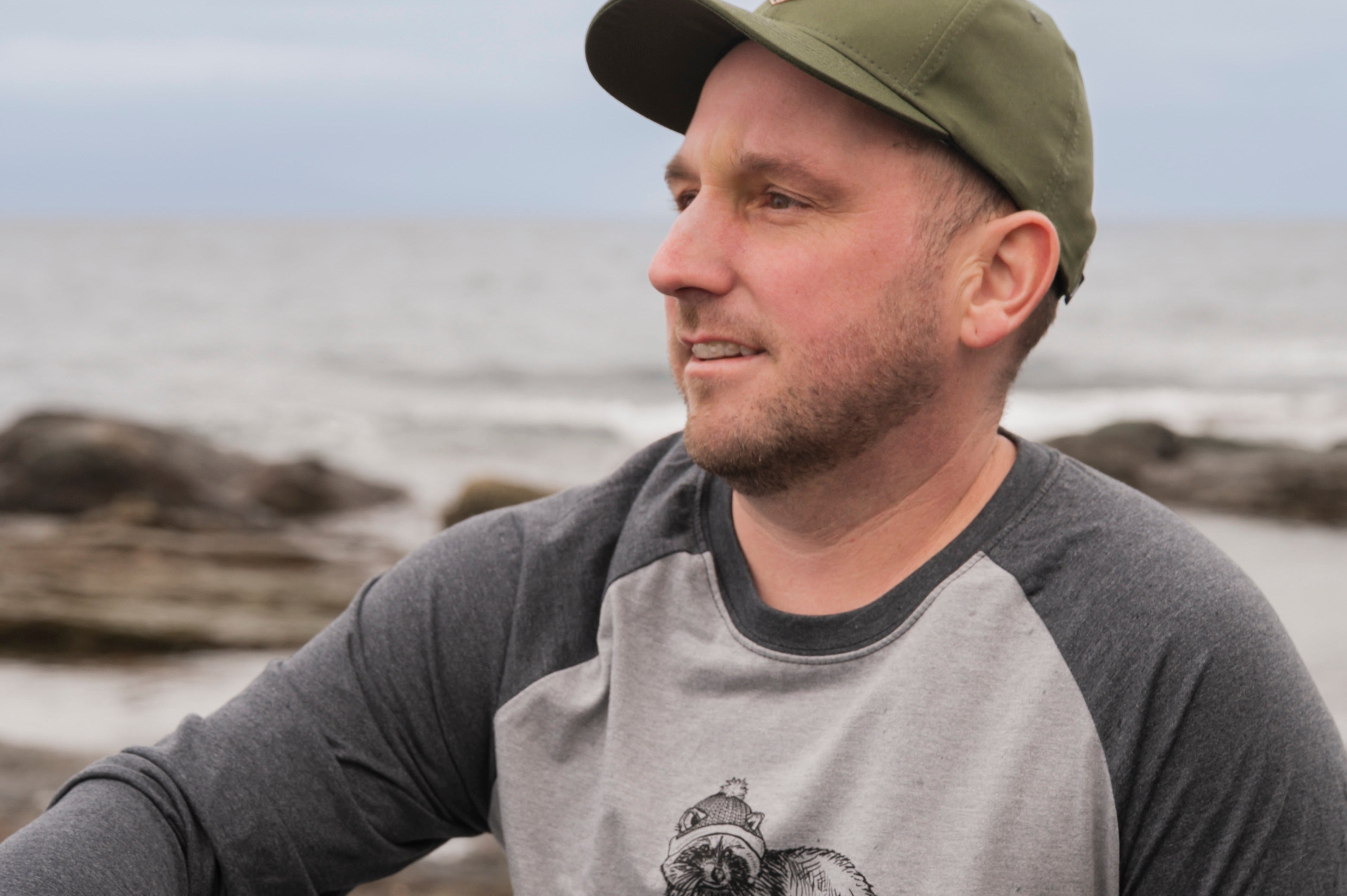 Man wearing a gray baseball shirt with a cat graphic sitting on a rocky beach.