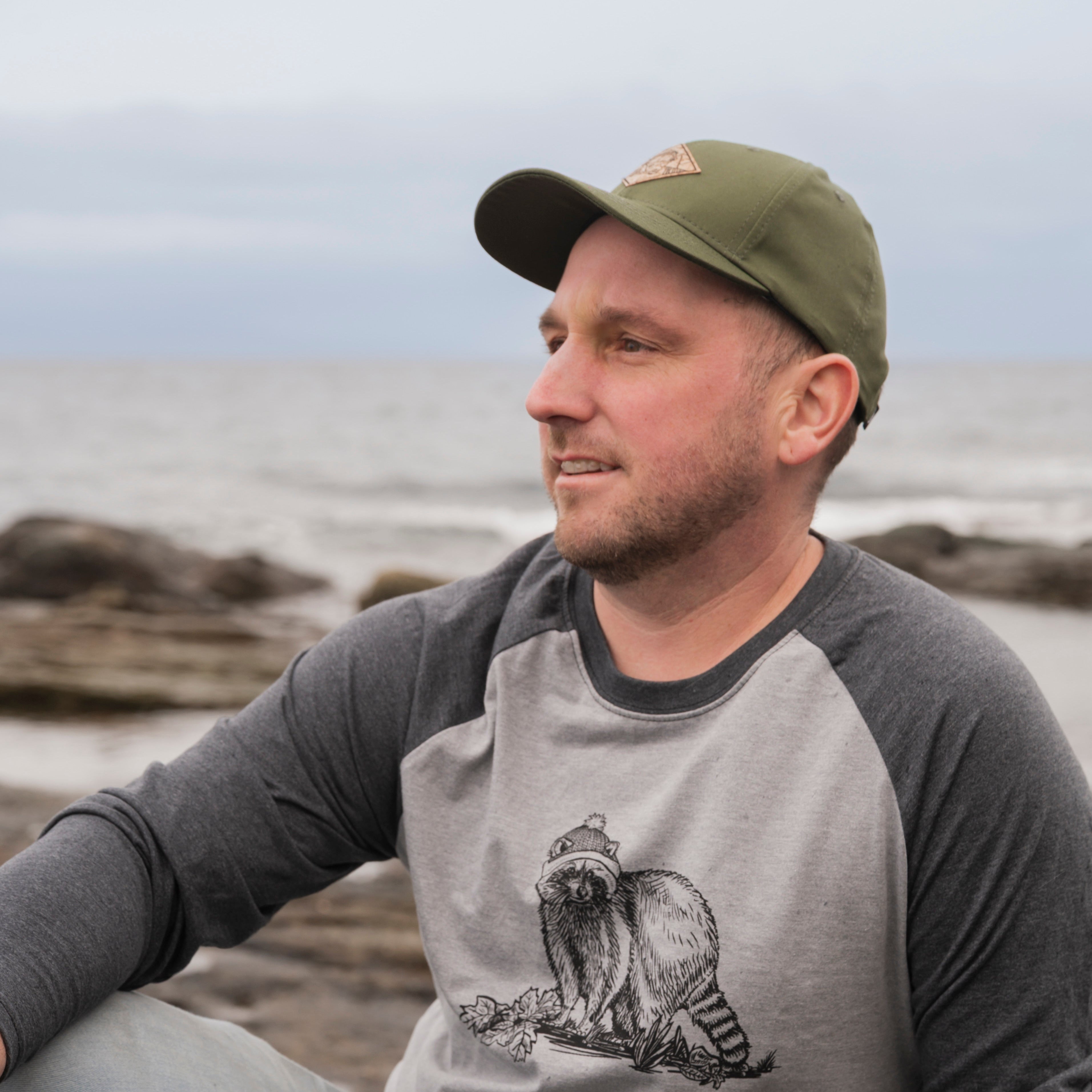 Man wearing a gray baseball shirt with a cat graphic sitting on a rocky beach.