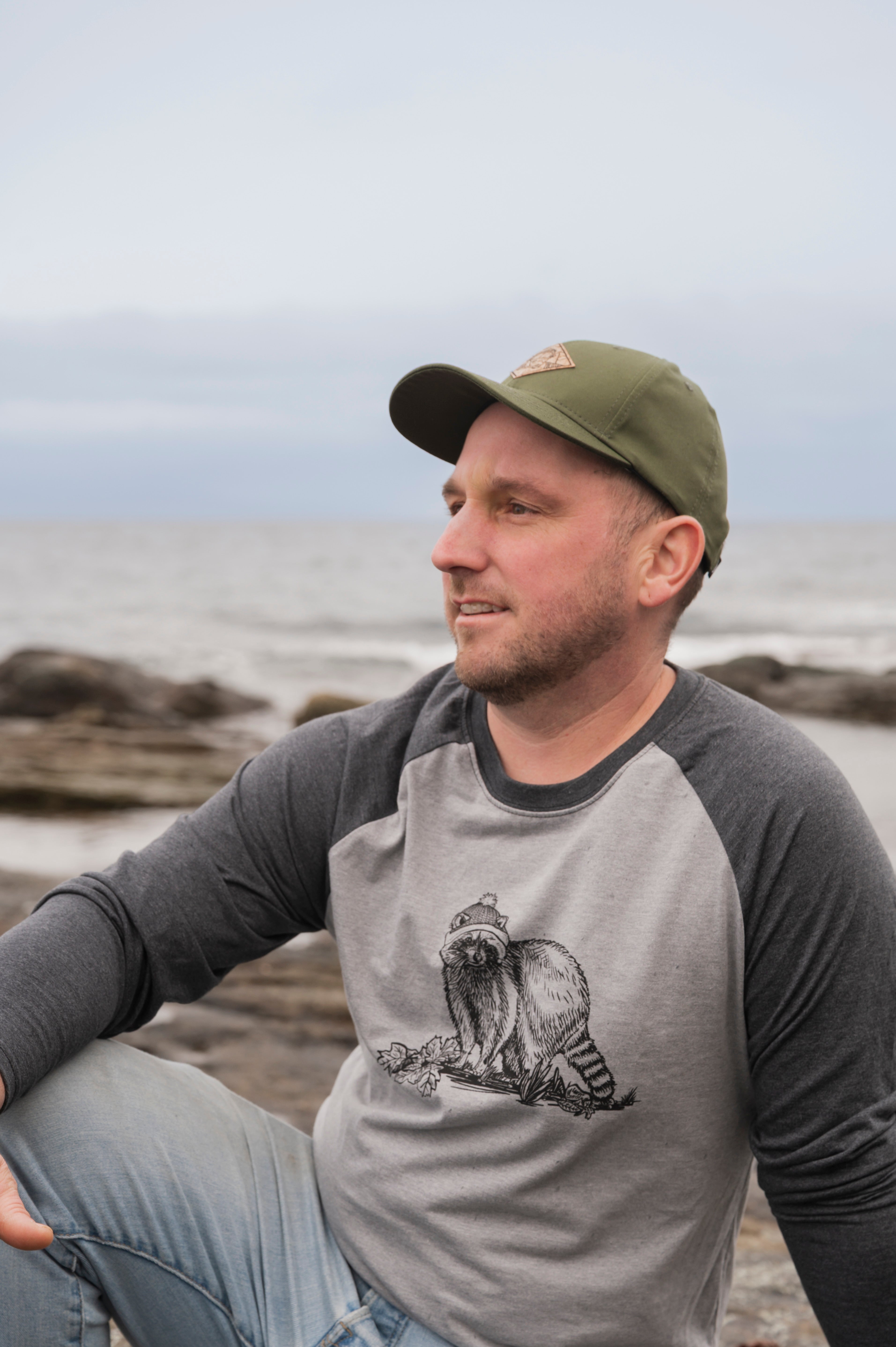 Man wearing a gray baseball shirt with a cat graphic sitting on a rocky beach.