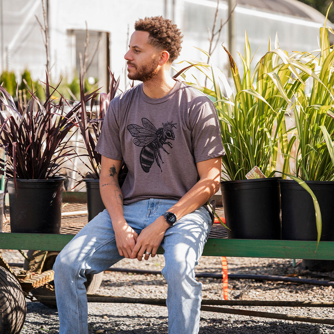 Man sitting on a bench in a garden setting with plants and a greenhouse in the background.