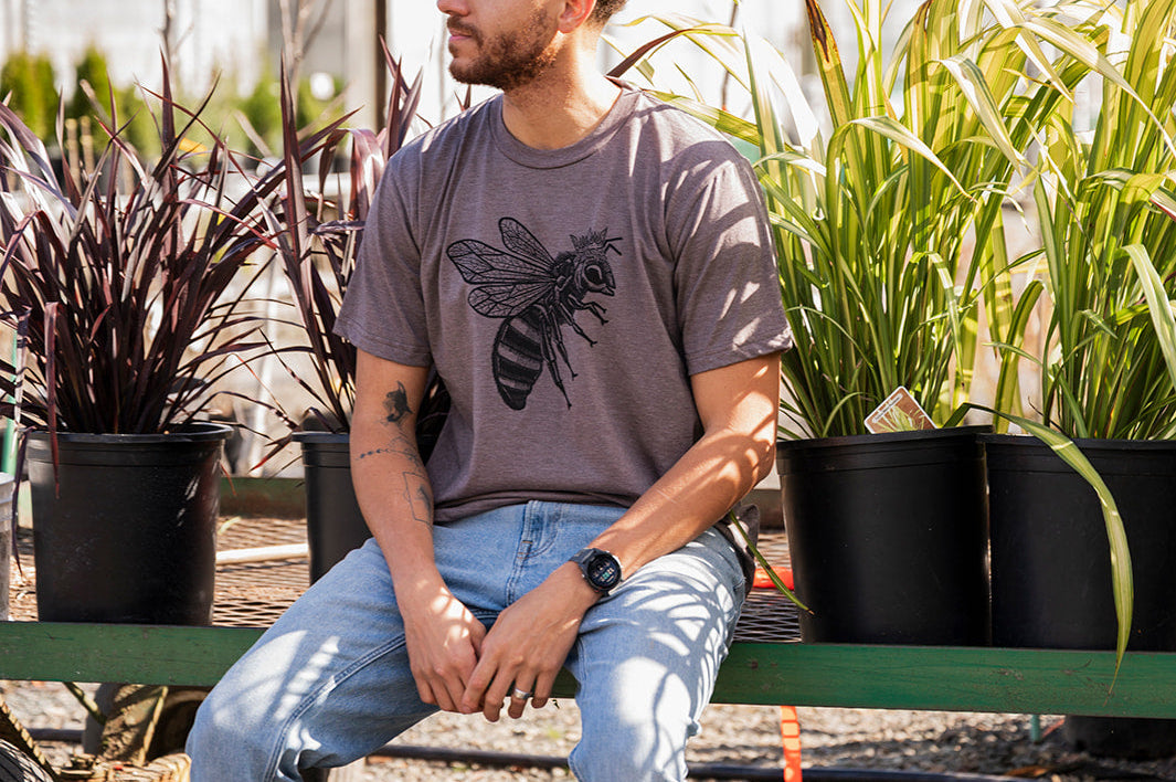 Man sitting on a bench in a garden setting with plants and a greenhouse in the background.
