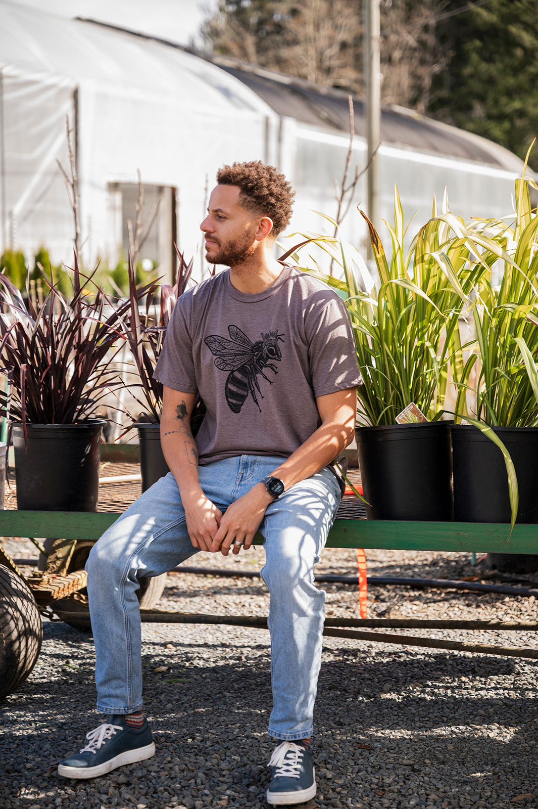 Man sitting on a bench in a garden setting with plants and a greenhouse in the background.