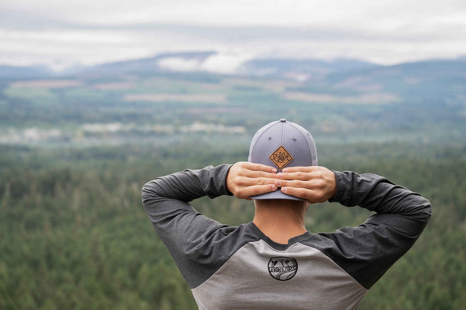 Person wearing a cap with a logo, standing with back to camera, overlooking a scenic landscape.