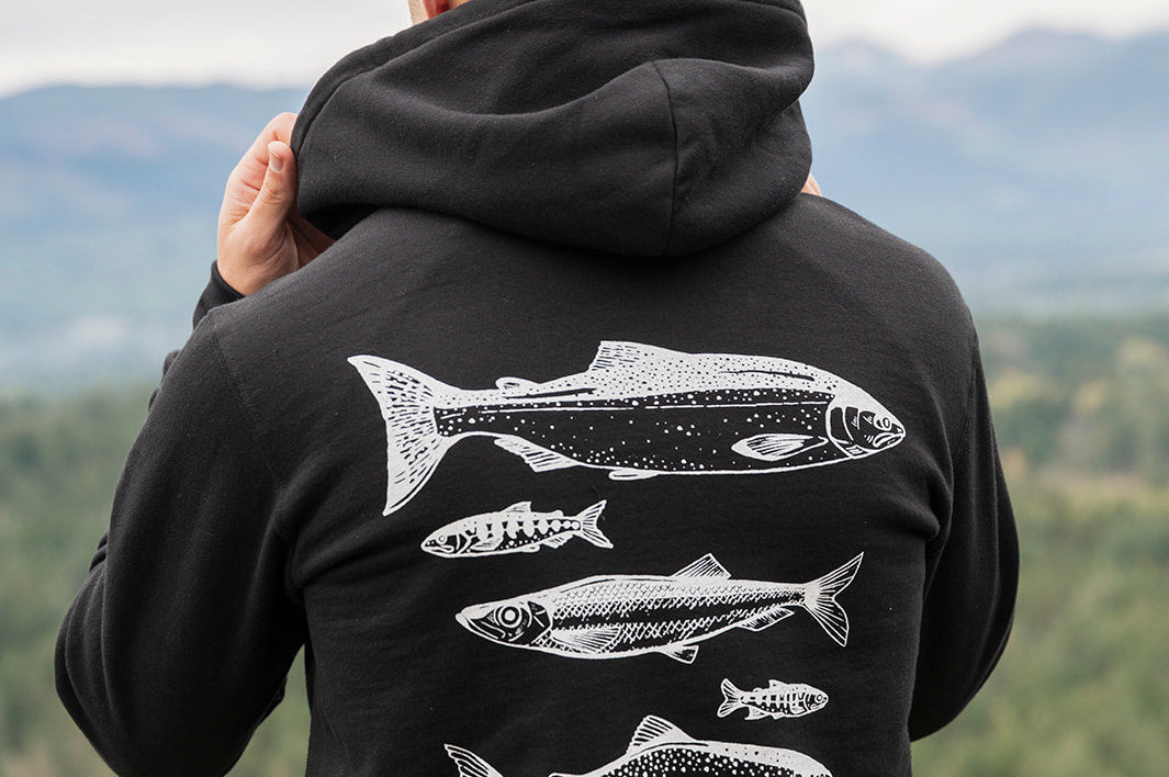 Person wearing a black hoodie with fish designs on the back, standing in a scenic outdoor setting.