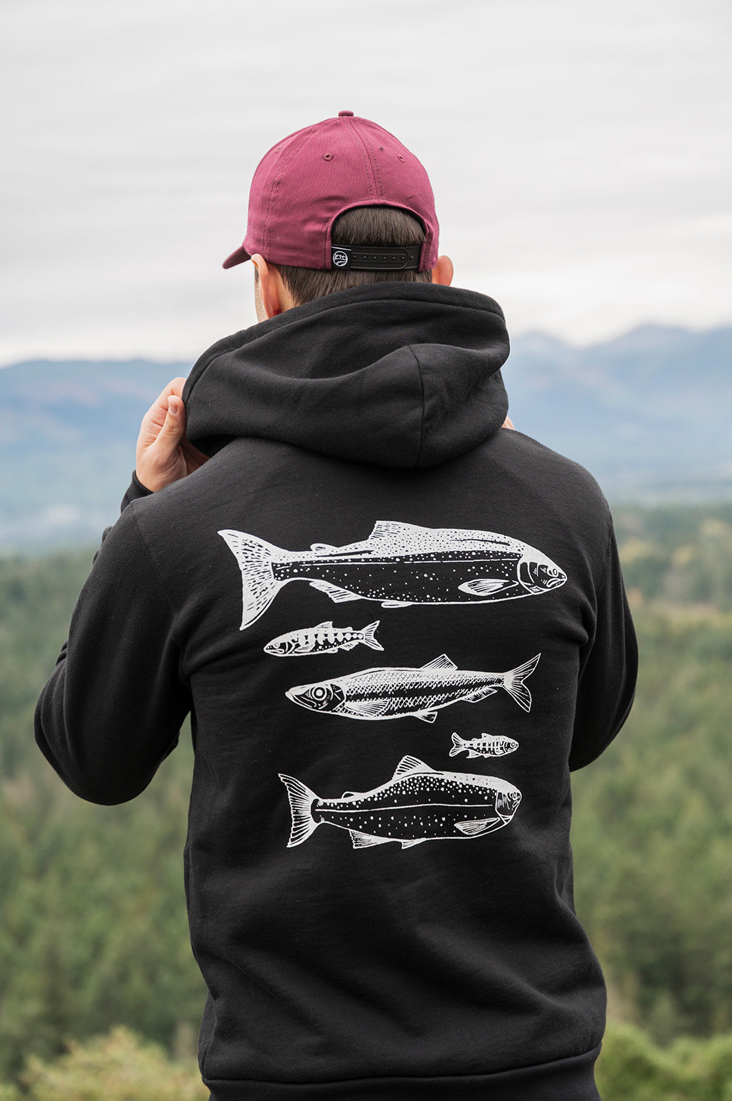 Person wearing a black hoodie with fish designs on the back, standing in a scenic outdoor setting.