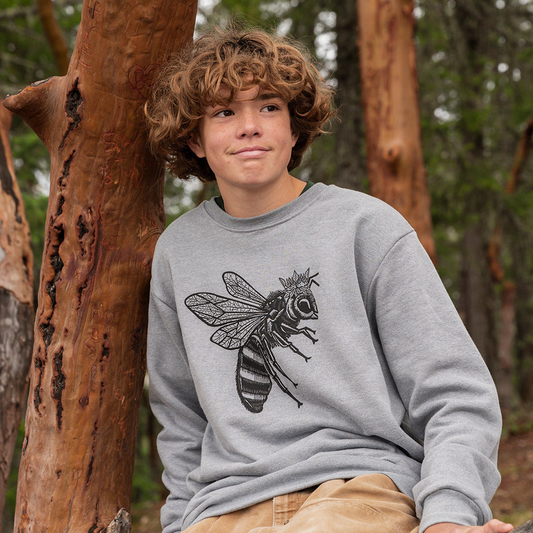 Person wearing a gray sweatshirt with a bee design, sitting against a tree in a forest.