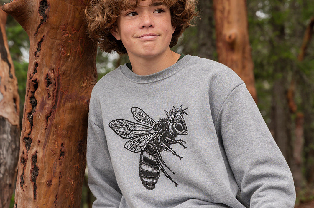 Person wearing a gray sweatshirt with a bee design, sitting against a tree in a forest.