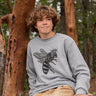 Person wearing a gray sweatshirt with a bee design, sitting against a tree in a forest.