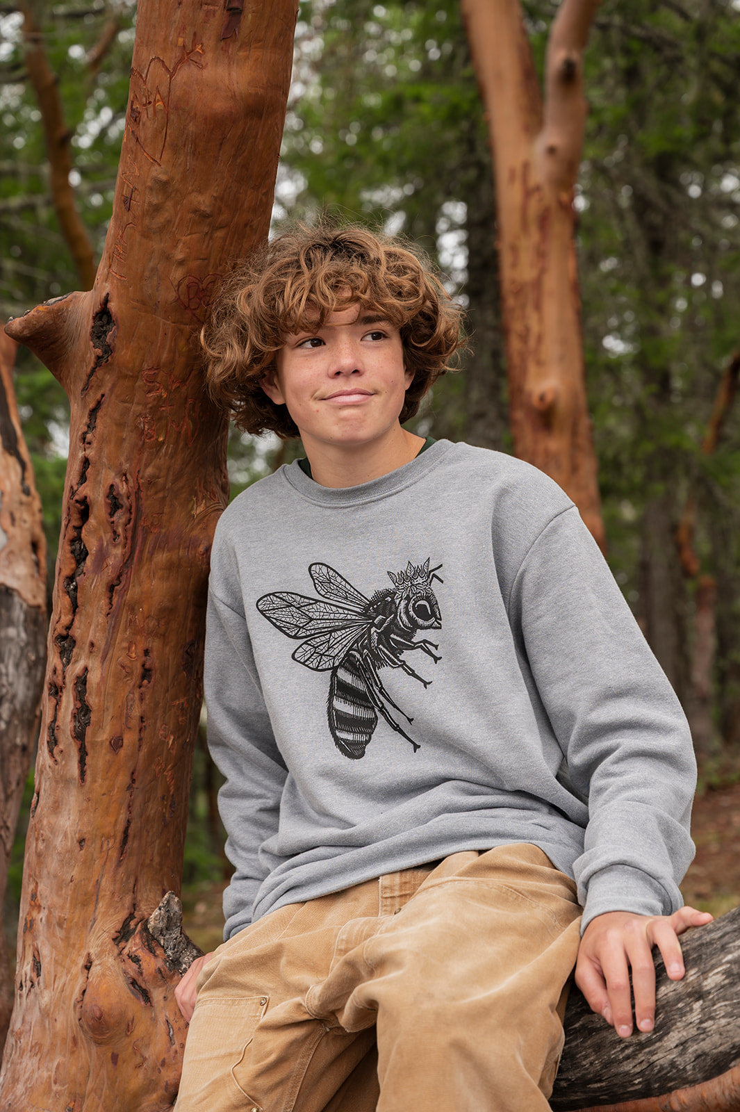 Person wearing a gray sweatshirt with a bee design, sitting against a tree in a forest.