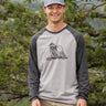 Man wearing a gray and black baseball shirt with a graphic design, standing in a forest.