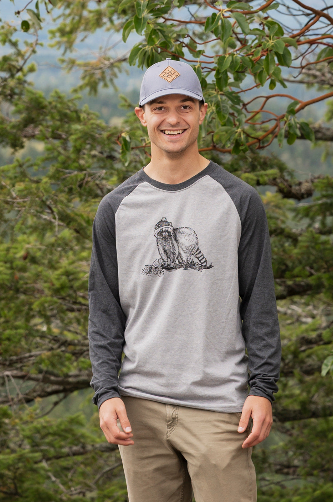 Man wearing a gray and black baseball shirt with a graphic design, standing in a forest.