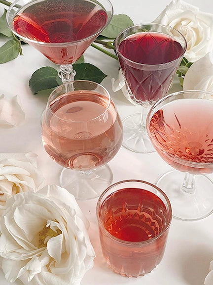 Assorted glasses of vibrant hibiscus tea drinks with white roses on a table, showcasing rose and fruity colors.