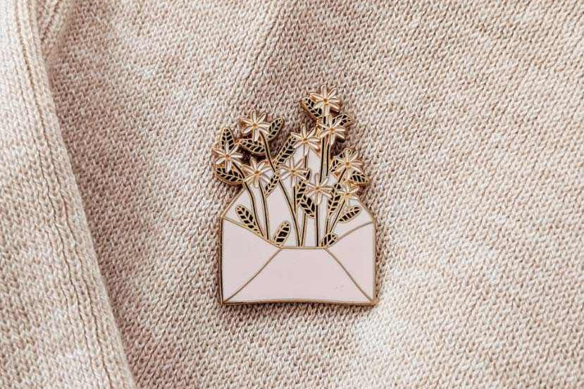 Enamel pin of a bouquet in an envelope on a beige fabric background mimi-and-august