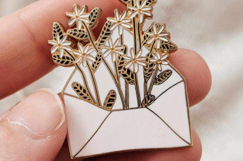 Pin with floral design held between fingers against a neutral background mimi-and-august