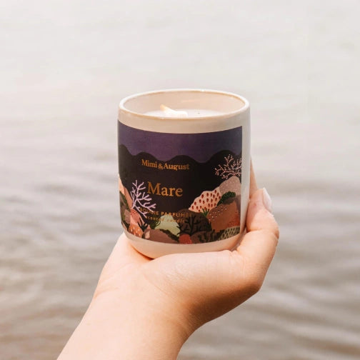 Hand holding a candle labeled 'Mare' by 'Mini & August' against a neutral background