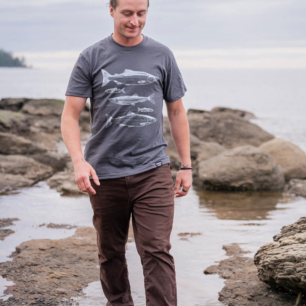 Man standing on a rocky shoreline wearing a gray t-shirt with a fish design.