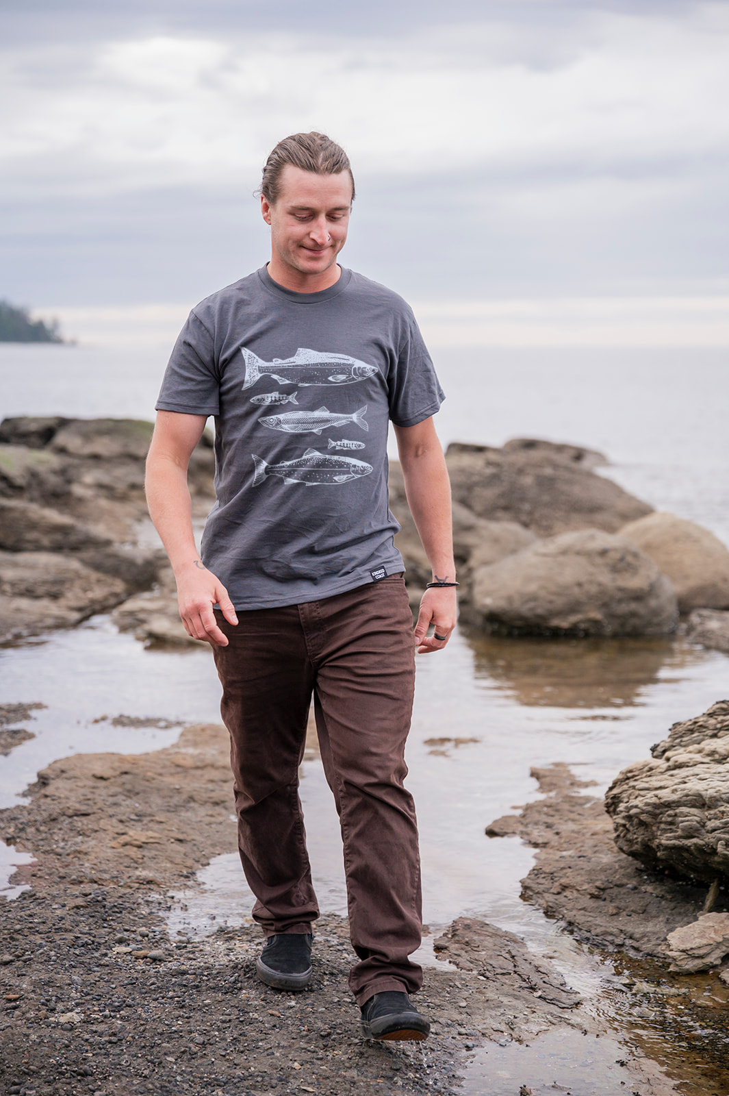 Man standing on a rocky shoreline wearing a gray t-shirt with a fish design.