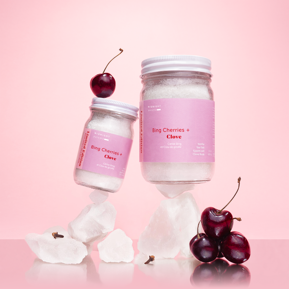 Bing Cherries + Clove Bath Soak jars with cherries on pink background, showcasing luxurious winter bath indulgence.