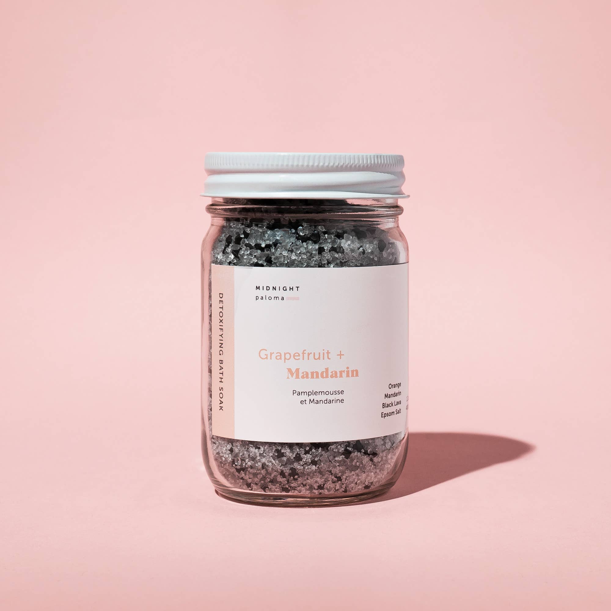Midnight Paloma Grapefruit + Mandarin Detoxifying Bath Soak in a jar with a simple label on a pink background.