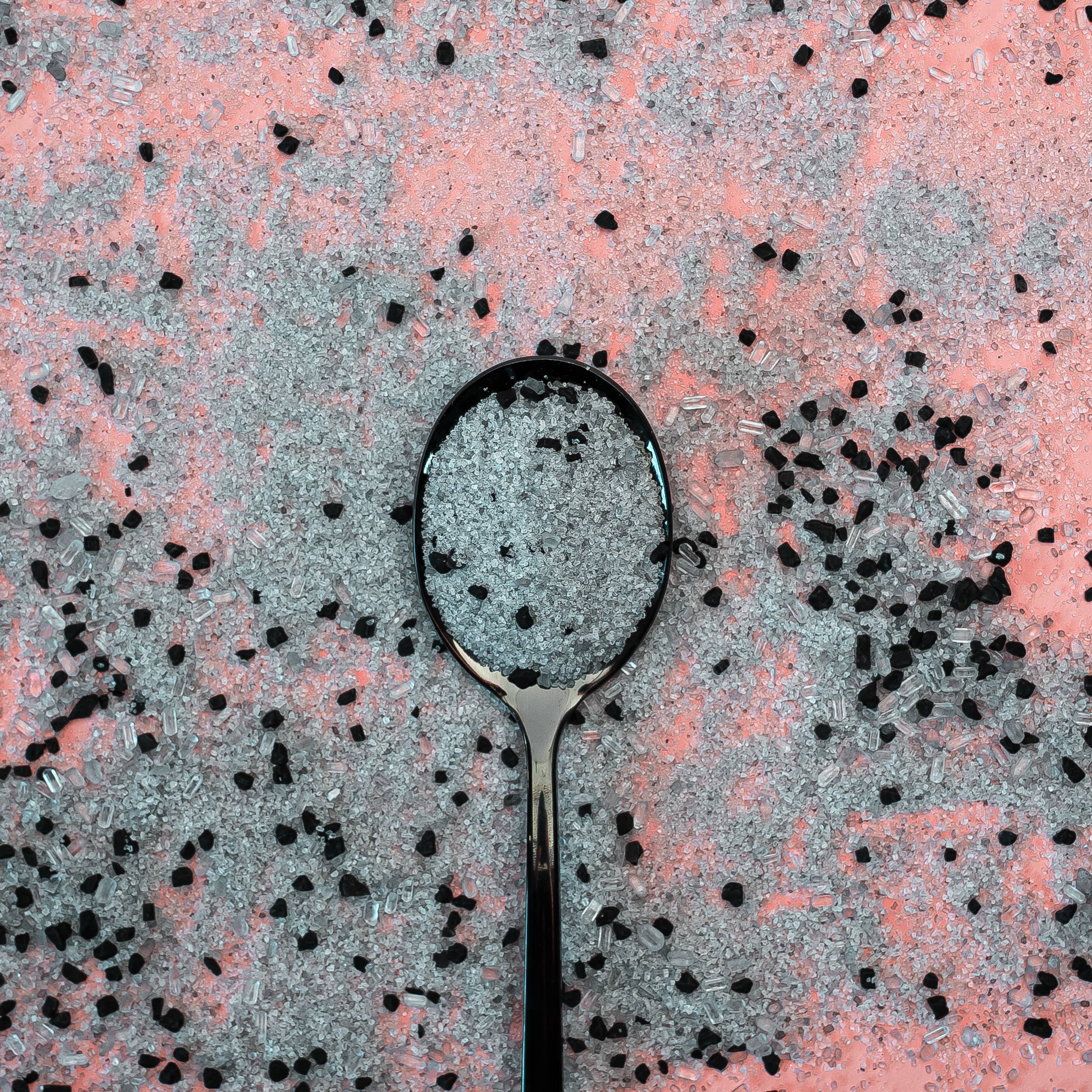 Close-up of a spoon surrounded by black and gray detoxifying bath salts on a pink background.