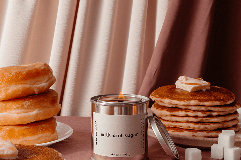 Candle labeled 'milk and sugar' surrounded by pancakes, donuts, and sugar cubes on a pink surface with curtains in the background.
