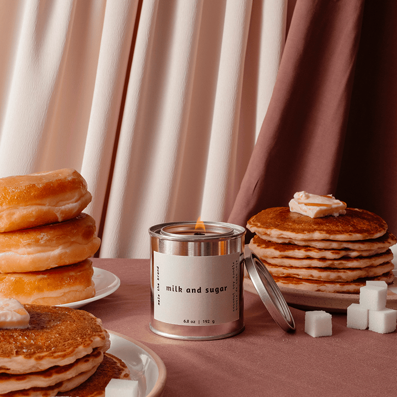 Candle labeled 'milk and sugar' surrounded by pancakes, donuts, and sugar cubes on a pink surface with curtains in the background.