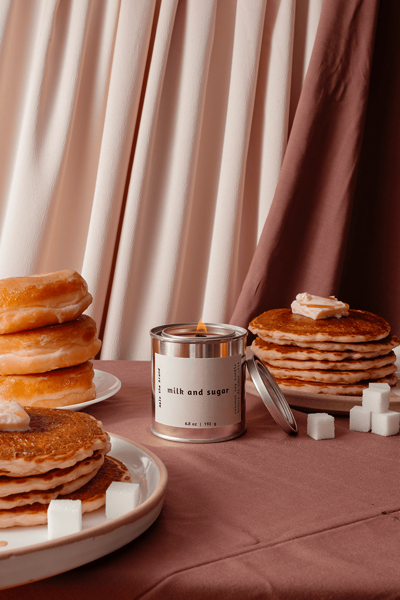 Candle labeled 'milk and sugar' surrounded by pancakes, donuts, and sugar cubes on a pink surface with curtains in the background.