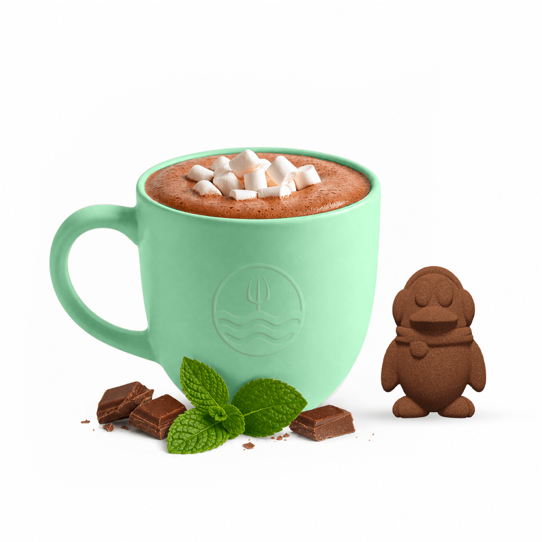 A mint green ceramic mug filled with hot chocolate and topped with marshmallows, styled with chocolate pieces, mint leaves, and a small gingerbread figure on a white background.