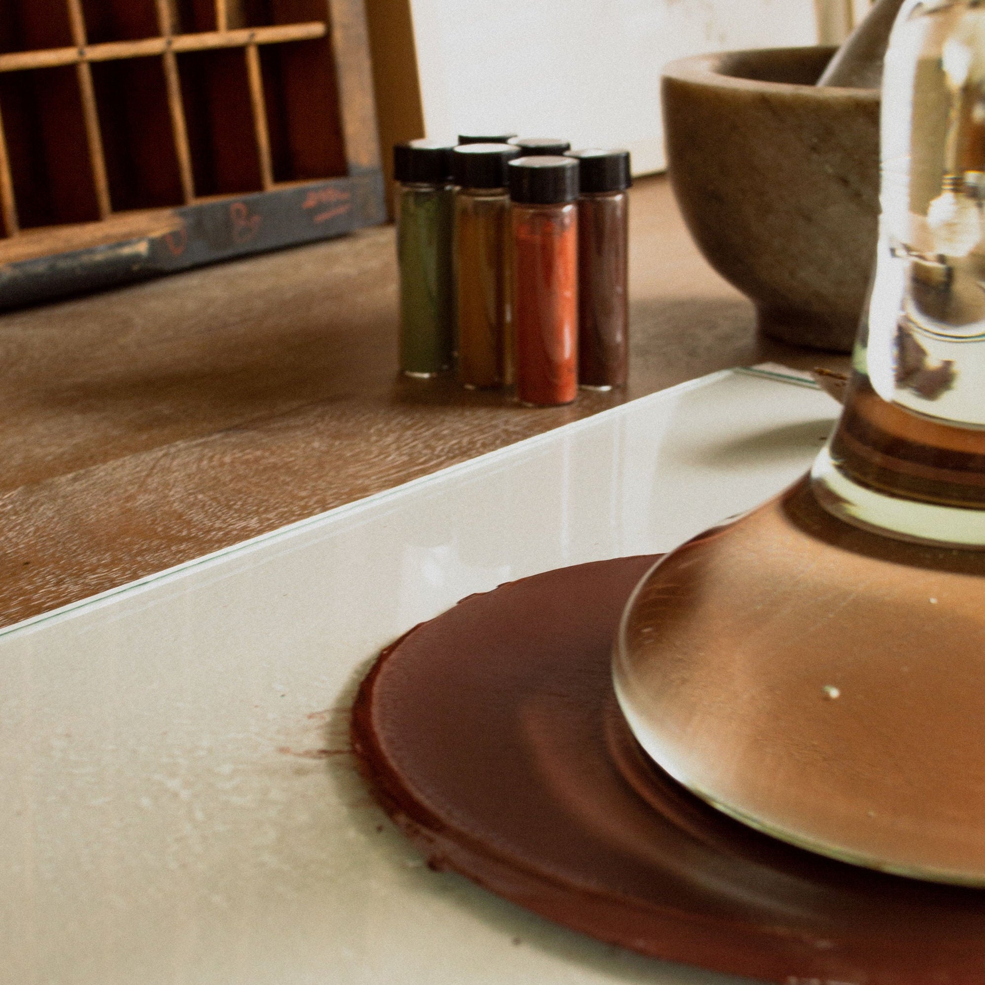 Clear Glass Paint Muller with a brown cap on a white surface, with a wooden shelf and decorative items in the background.