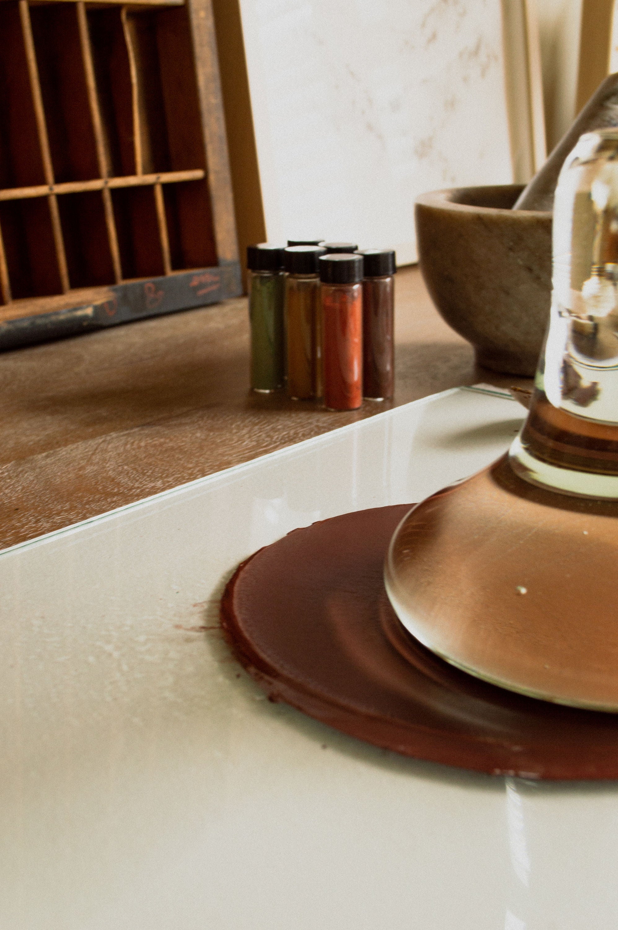Clear Glass Paint Muller with a brown cap on a white surface, with a wooden shelf and decorative items in the background.