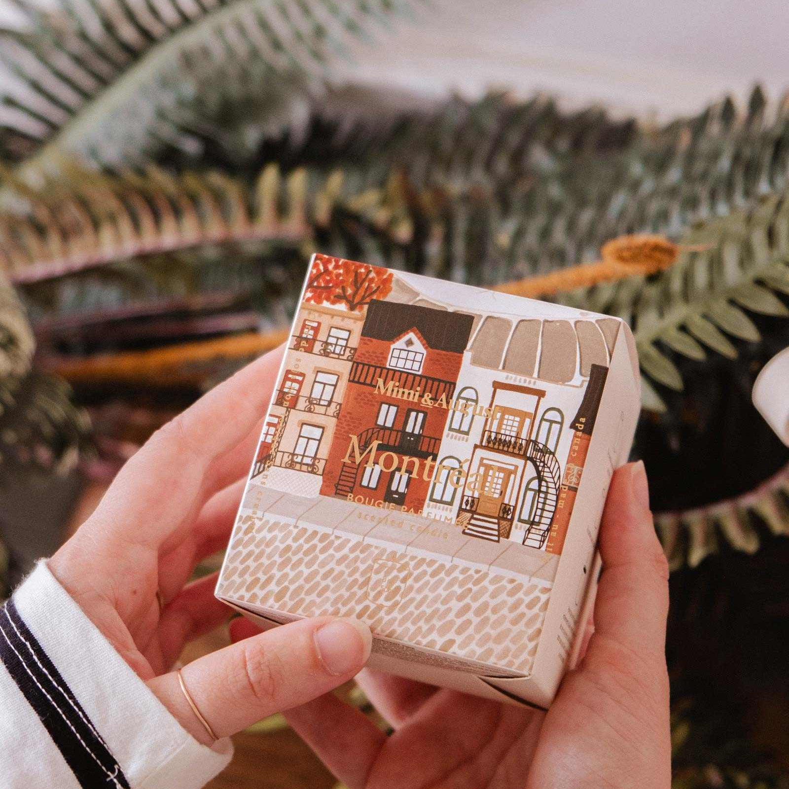 Hand holding a small box with a decorative design of houses, surrounded by green foliage.