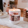 Candle in a decorative mug with a town design on a marble surface.