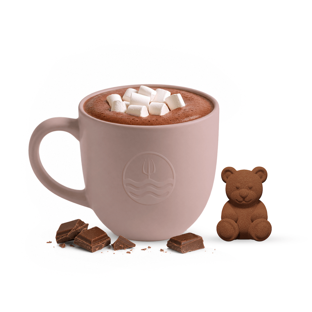 A dusty pink ceramic mug filled with hot chocolate and topped with mini marshmallows, shown on a white background with chocolate chunks and a small chocolate teddy bear beside it.