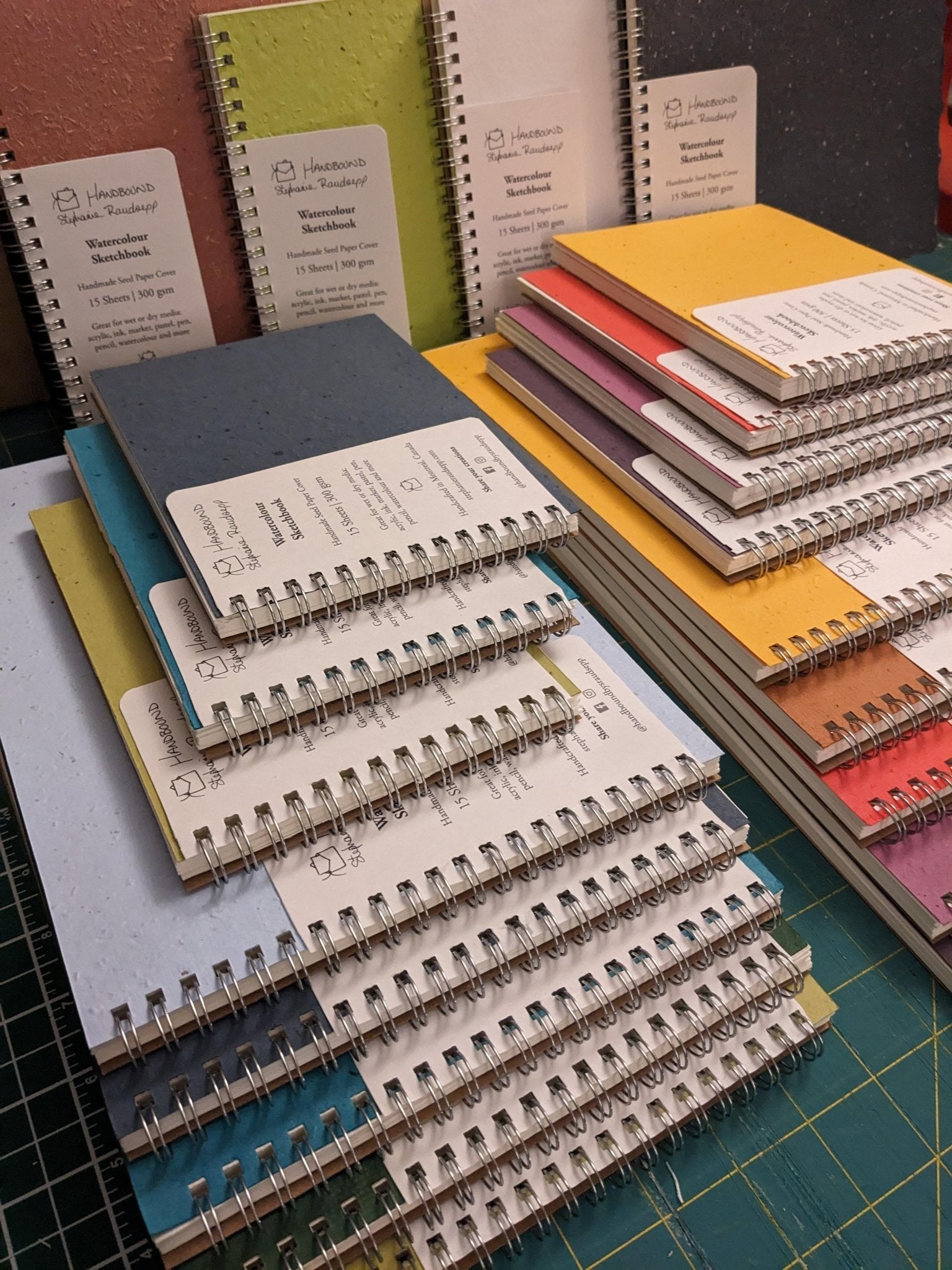 Stacks of colorful notebooks on a table with a cutting mat underneath Plantable Watercolour Sketchbook by Creative Artifacts HQ